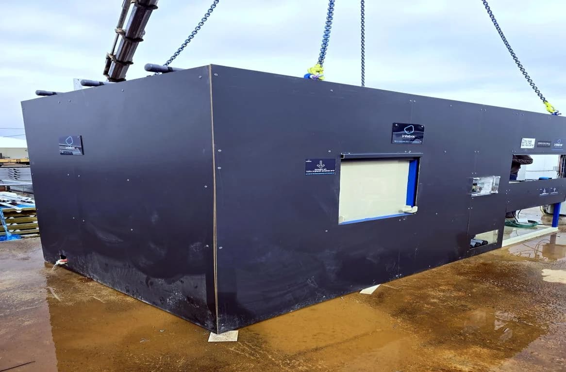A hi-vis crew member watching a black INFRABOX unit being lowered from a crane onto its pad beside a water tank.
