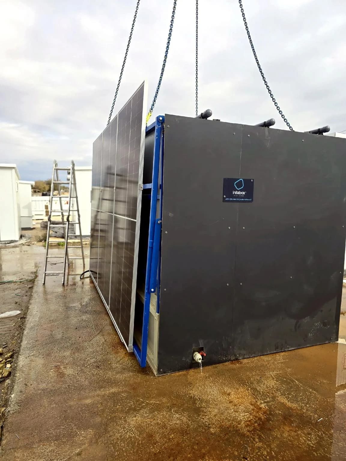 A matte black INFRABOX unit on crane chains above a wet concrete pad, its solar array folded against the blue lifting frame and a ladder standing beside it