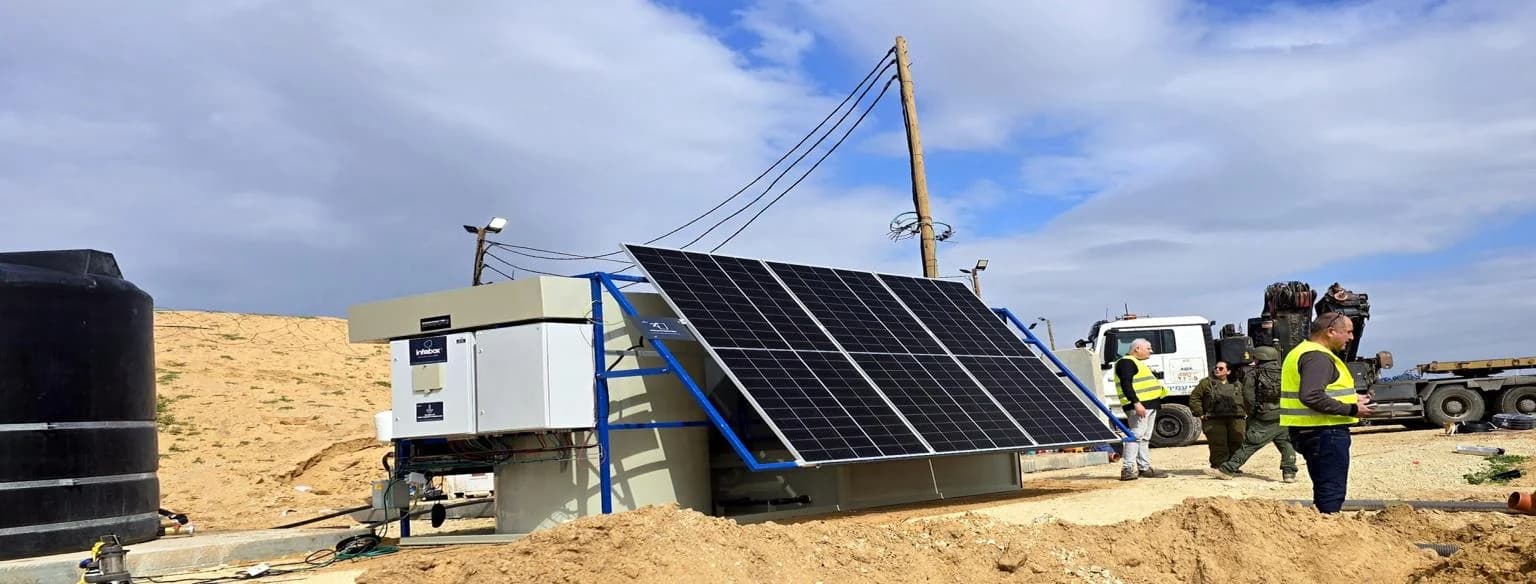 Une unité de traitement INFRABOX, son ensemble de panneaux solaires à cadres bleus et la dalle en béton sur un site désertique nivelé, avec des lignes aériennes à l’arrière-plan.