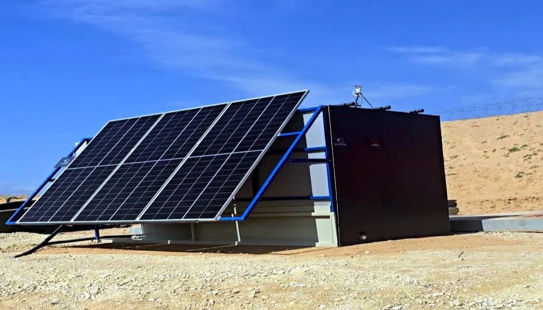 A blue-framed solar array tilted toward the sun beside a black INFRABOX treatment unit on a cut desert pad.