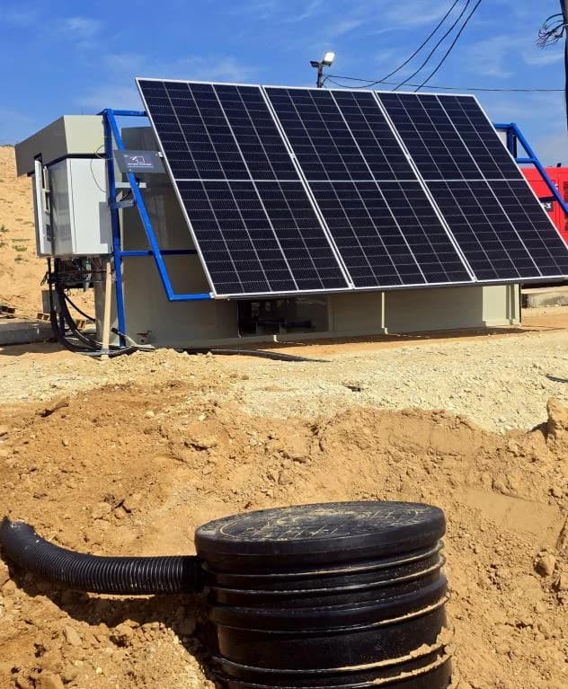 A blue-framed solar array tilted over an INFRABOX unit on a sand pad, with the site's power line running overhead.