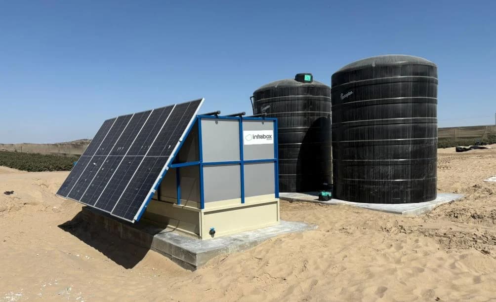 A black INFRABOX unit with its solar panel leaning against it and two black water tanks on concrete pads in open sand.