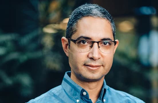 In glasses and a blue denim shirt, photographed against a dark interior with planting behind.