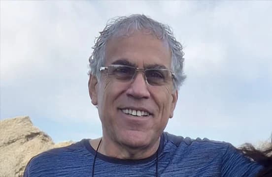 شعر رمادي، بنظارة بلا إطار وقميص أزرق مضلّع، مصوَّر في الخارج أمام صخر فاتح وسماء مفتوحة.
